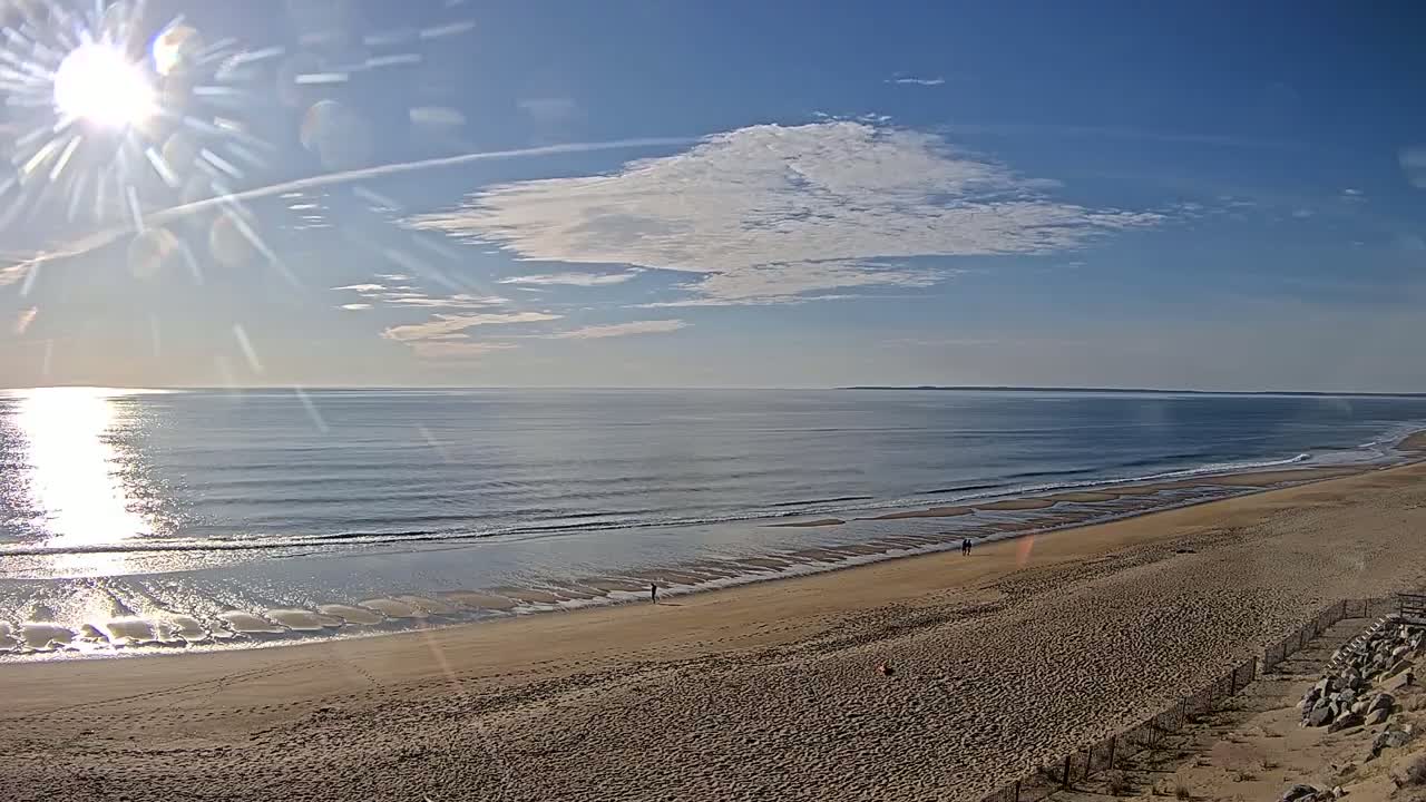 A sandy beach stretches along a calm ocean under a mostly sunny sky with a few scattered clouds.