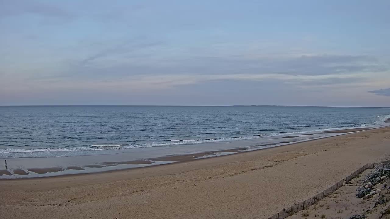 A long, sandy beach stretches toward a calm ocean under a mostly cloudy sky.