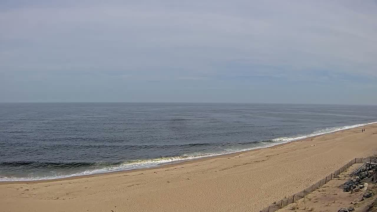 A long, sandy beach slopes down to a calm, dark-blue ocean under a mostly cloudy sky.