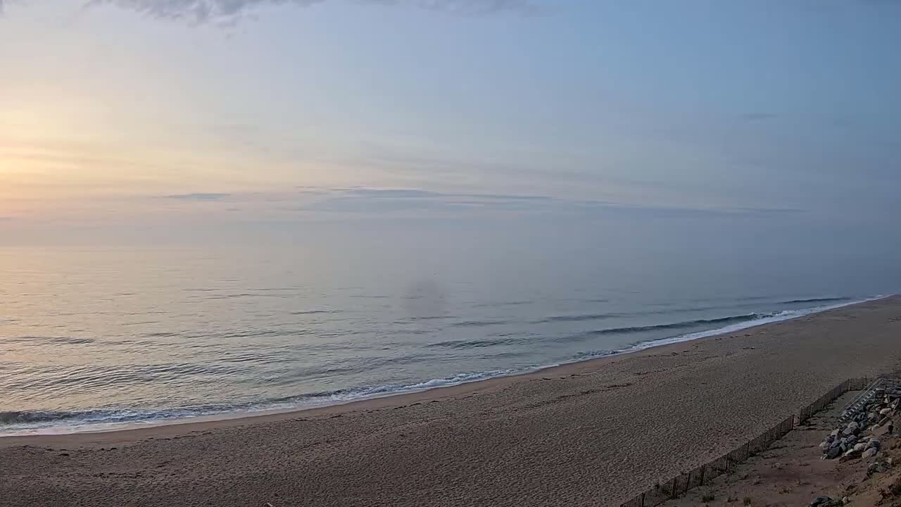 A sandy beach slopes down to a calm ocean under a hazy, sunrise sky.