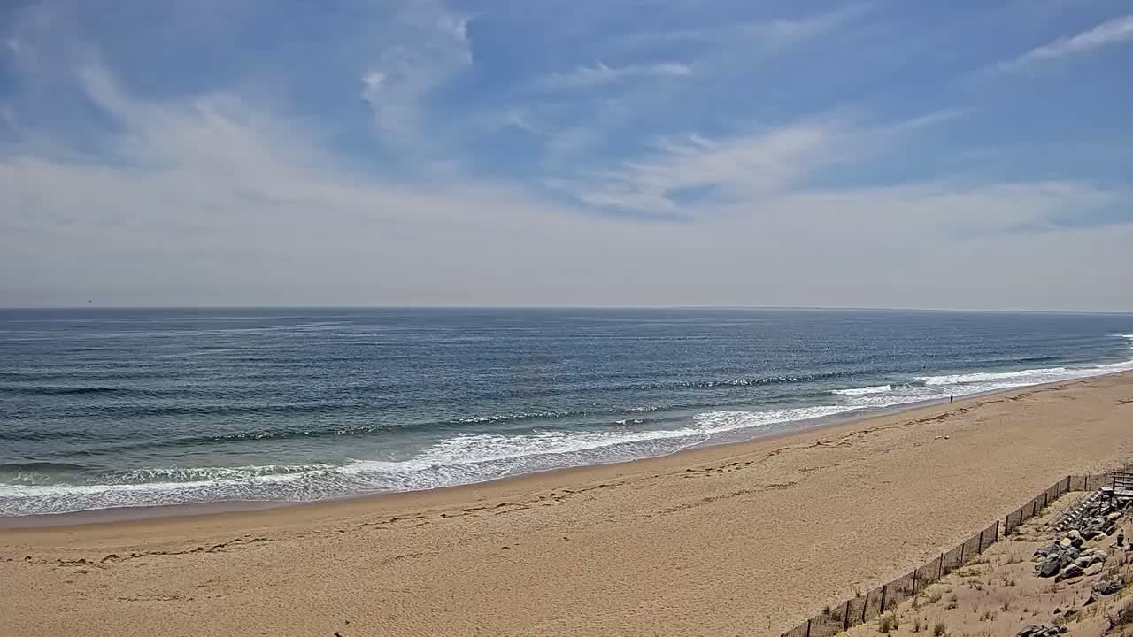 A wide sandy beach stretches along a calm ocean under a mostly sunny sky with scattered clouds.