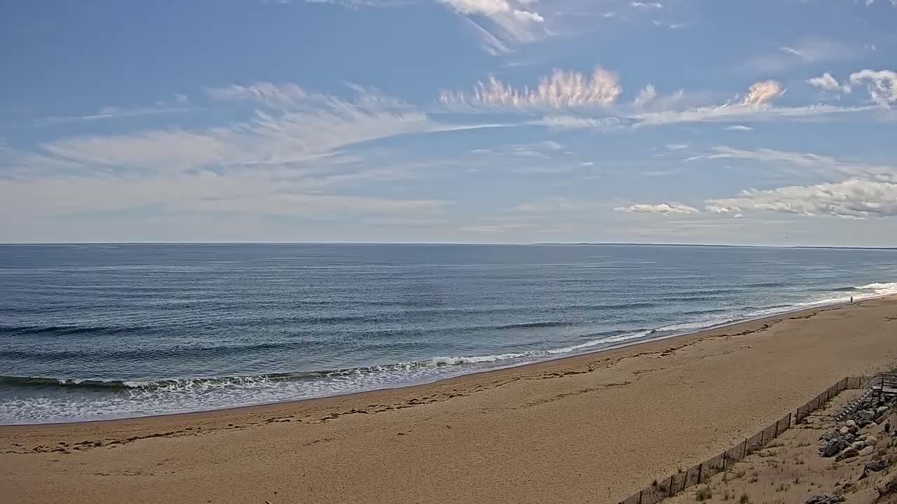 A sandy beach slopes gently down to a calm ocean under a mostly sunny sky with wispy clouds.