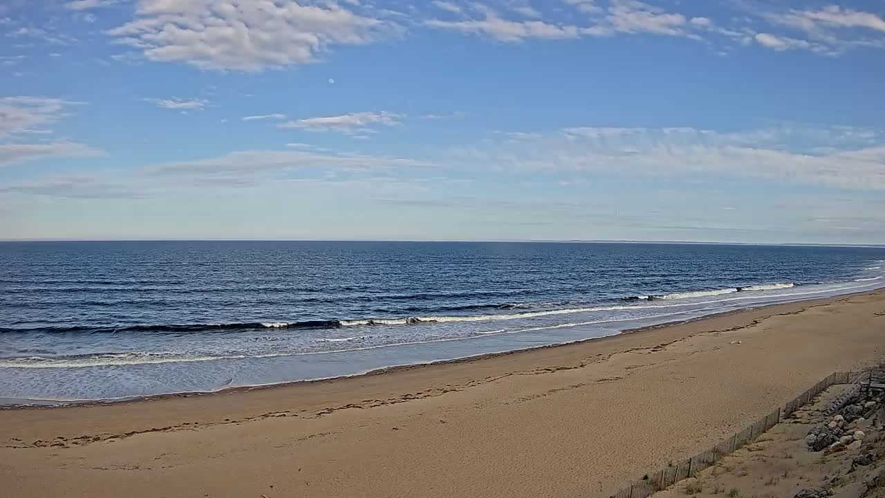 A sandy beach stretches along a calm ocean under a mostly clear sky with scattered clouds.