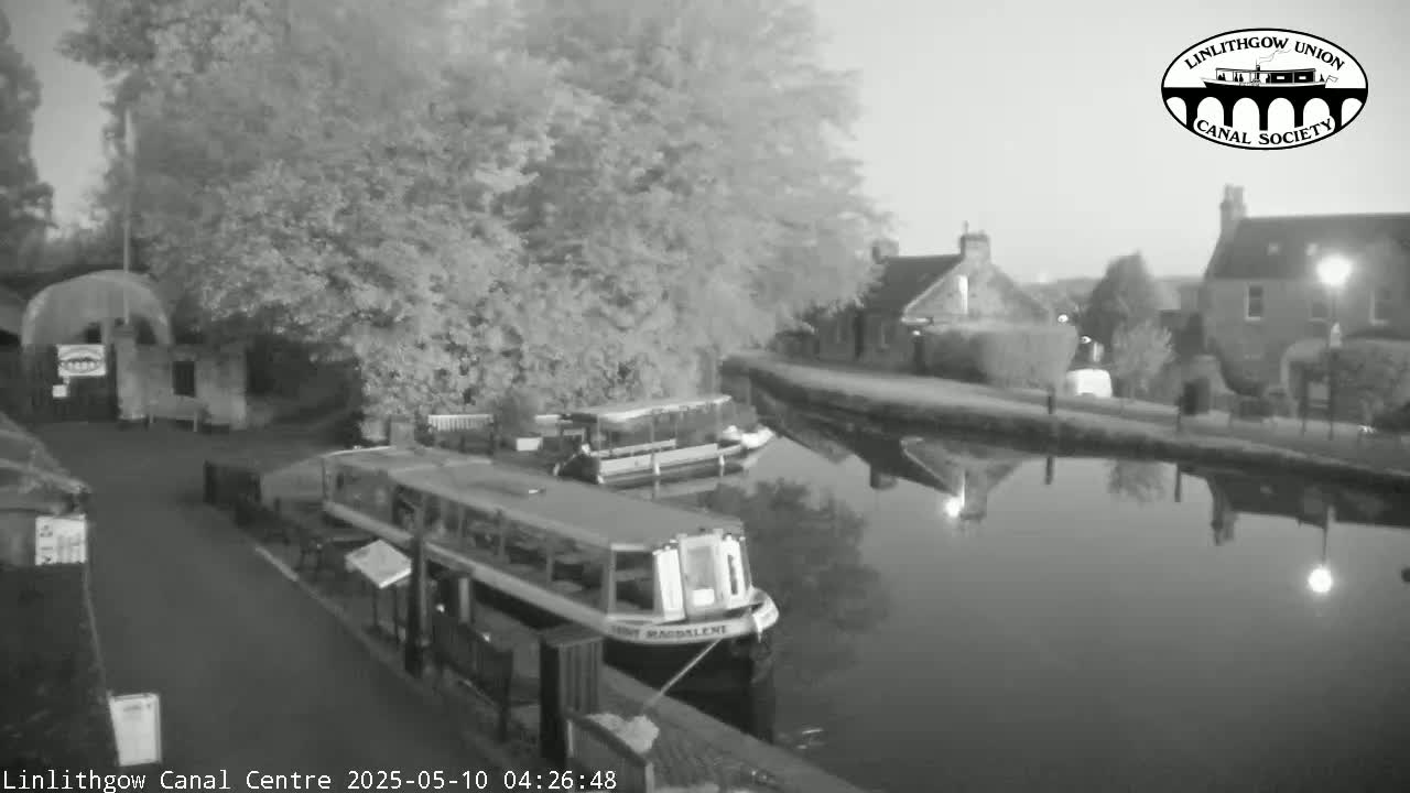 Two boats are moored on a calm canal at twilight, beside buildings and trees.