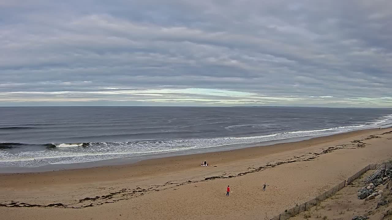 A sandy beach with small waves under a cloudy sky, a few people are visible on the beach.