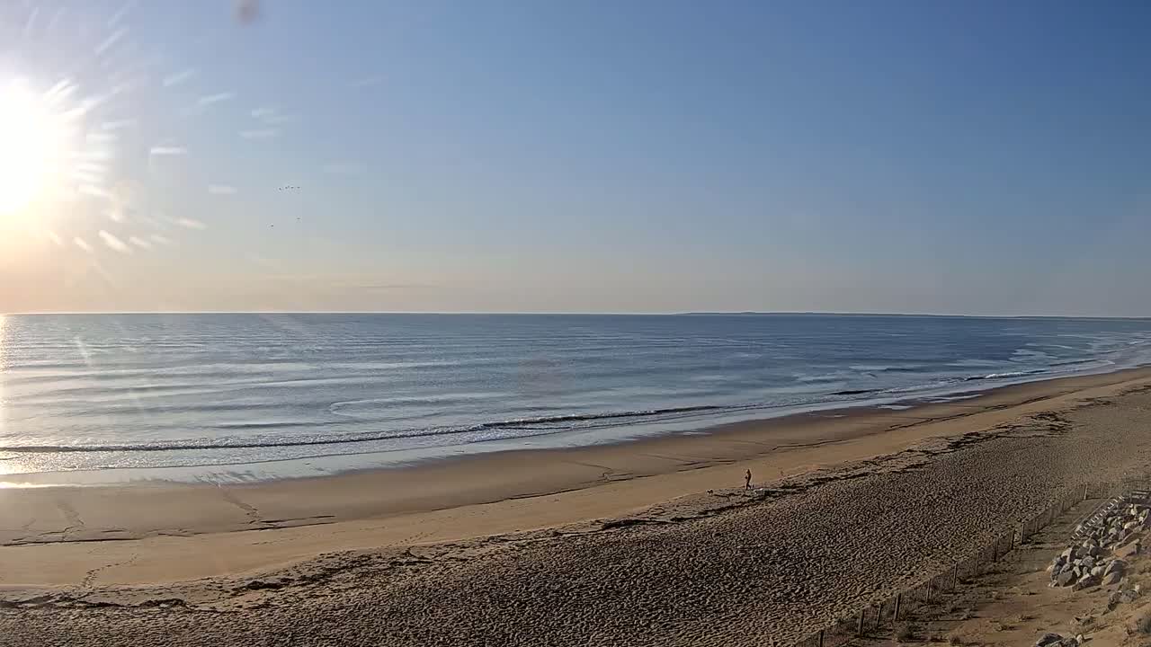 A sandy beach stretches along a calm ocean under a clear, sunny sky.