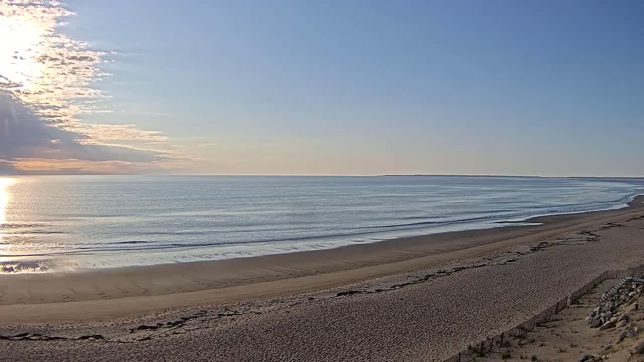 A sandy beach slopes gently down to a calm ocean under a partly cloudy sunrise sky.