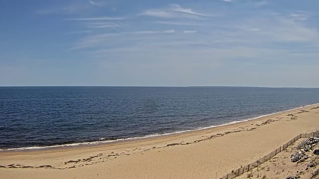 A sandy beach curves gently into a calm, dark-blue ocean under a mostly sunny sky with a few scattered clouds.