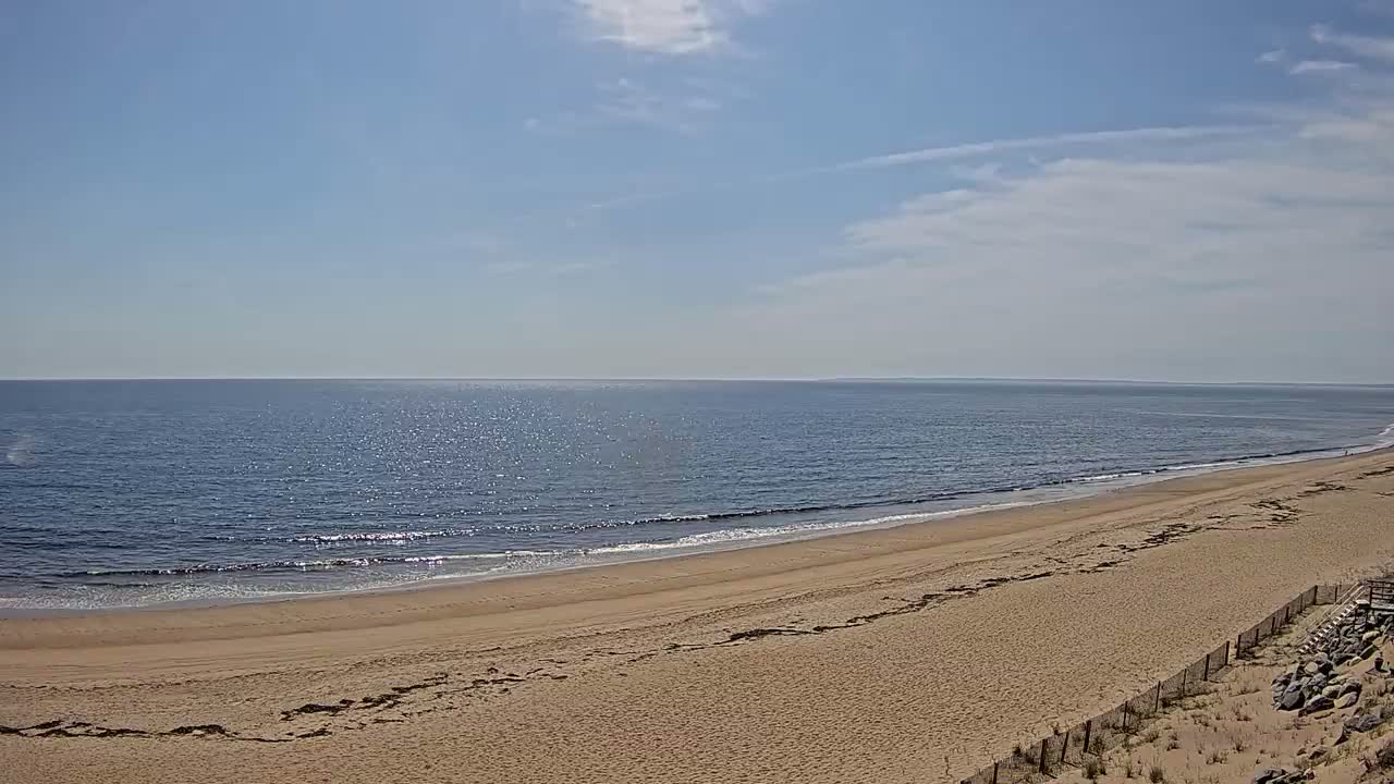 A long, sandy beach slopes gently down to a calm, sparkling ocean under a mostly sunny sky with a few scattered clouds.