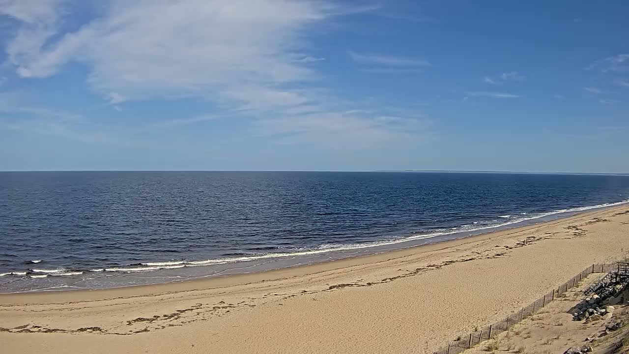 A sandy beach slopes gently down to a calm, blue ocean under a mostly sunny sky with a few scattered clouds.