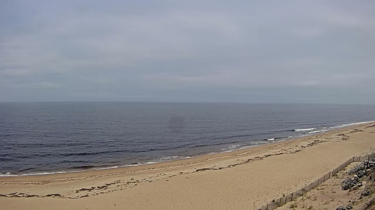 A long, sandy beach slopes gently down to a calm, dark-blue ocean under a mostly cloudy sky.