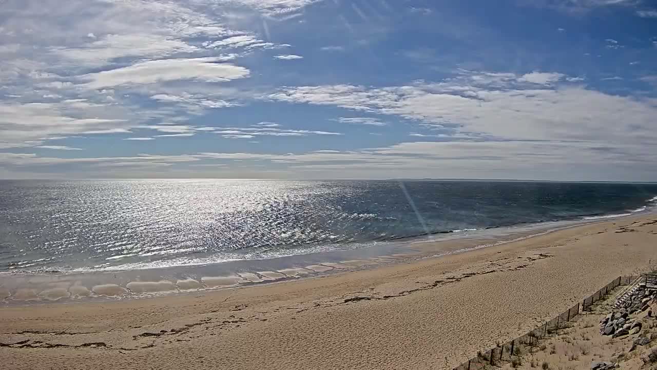 A sandy beach slopes down to a gently wavy ocean under a partly cloudy, sunny sky.