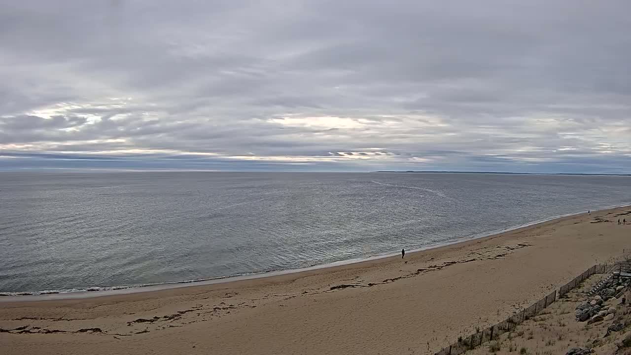 A sandy beach stretches along a calm ocean under a mostly cloudy sky.