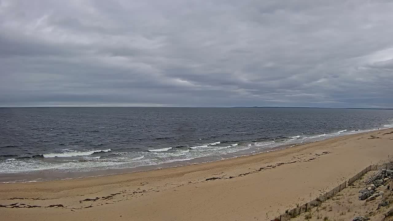 A wide sandy beach slopes down to a dark-blue ocean with small waves under a cloudy sky.