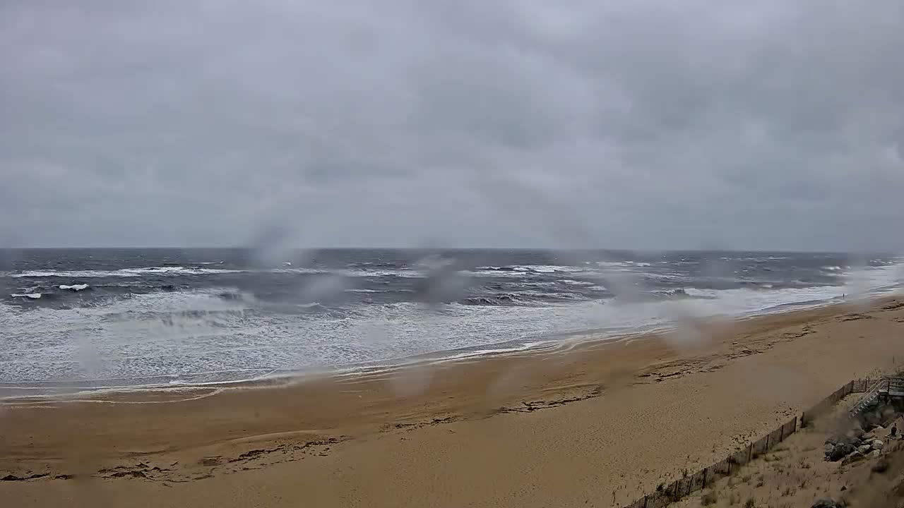 A wide sandy beach with choppy waves crashing on the shore under a gray, overcast sky with rain.