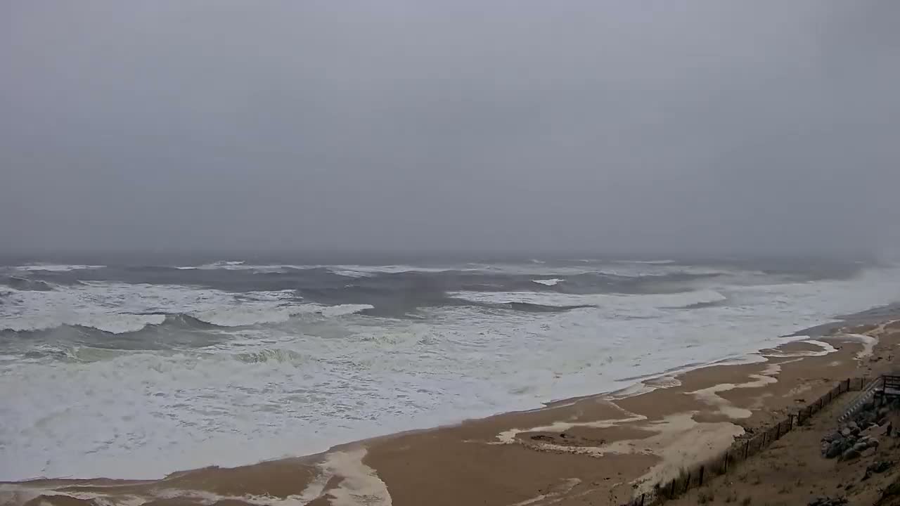 Large waves crash on a sandy beach under an overcast, gray sky.
