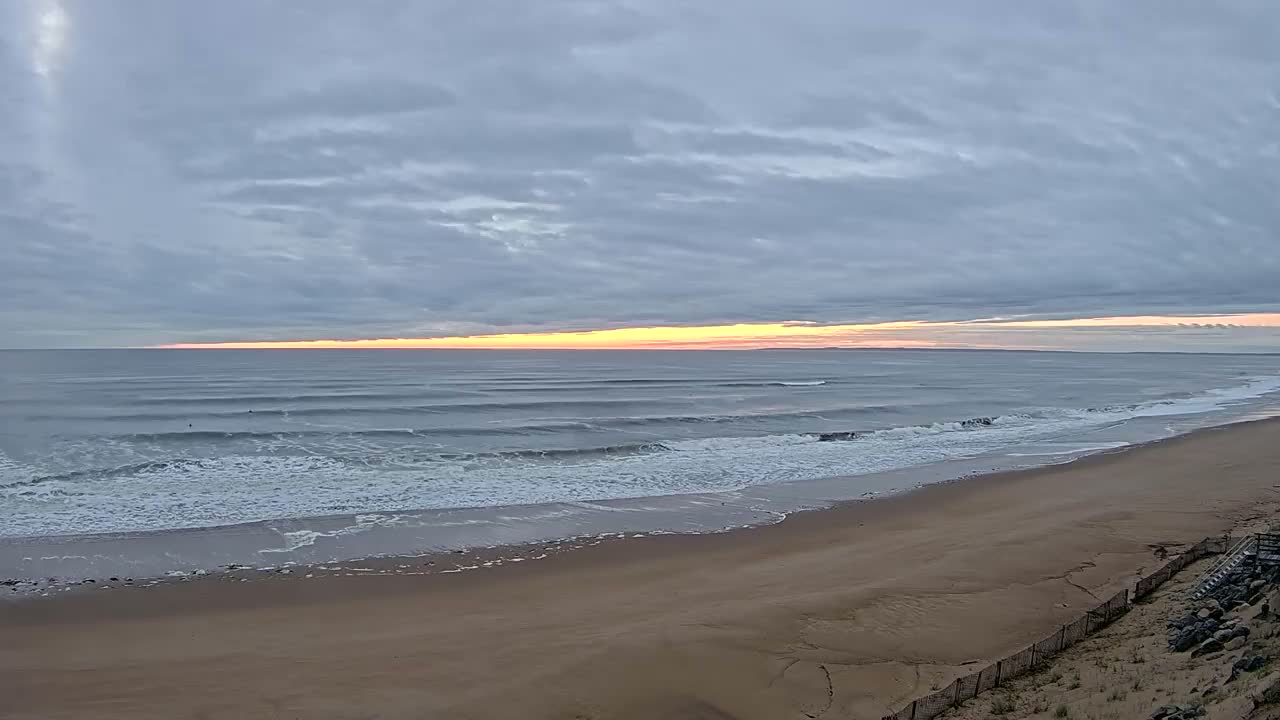 A long sandy beach stretches toward a calm ocean under a cloudy sky with a hint of sunrise on the horizon.