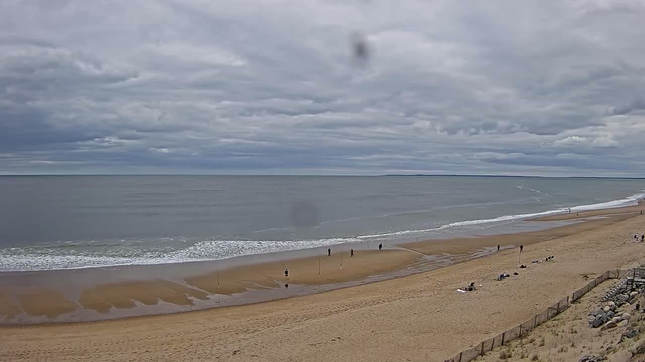 A wide sandy beach stretches to a calm, grey ocean under a cloudy sky, with a few people scattered along the shoreline.