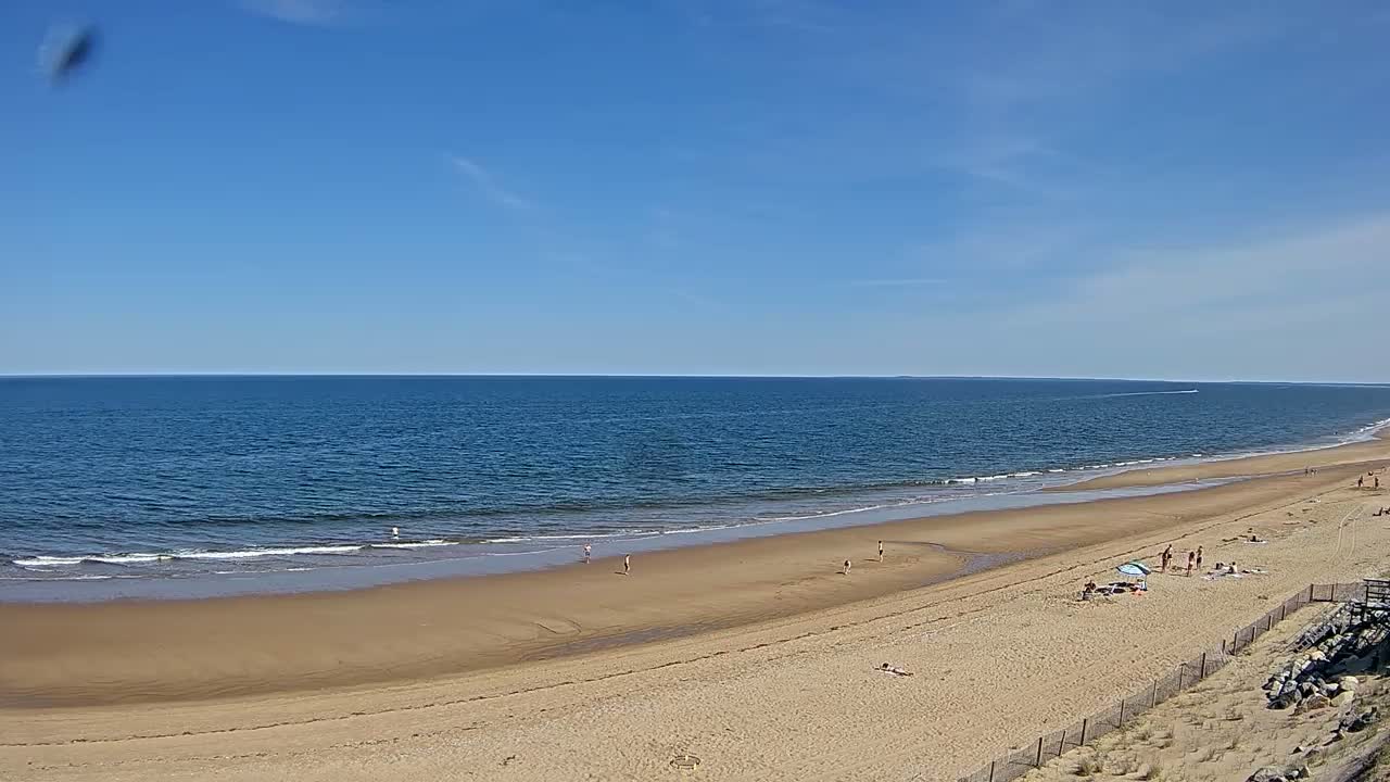 A sandy beach with a few people on it stretches along a calm ocean under a clear blue sky.