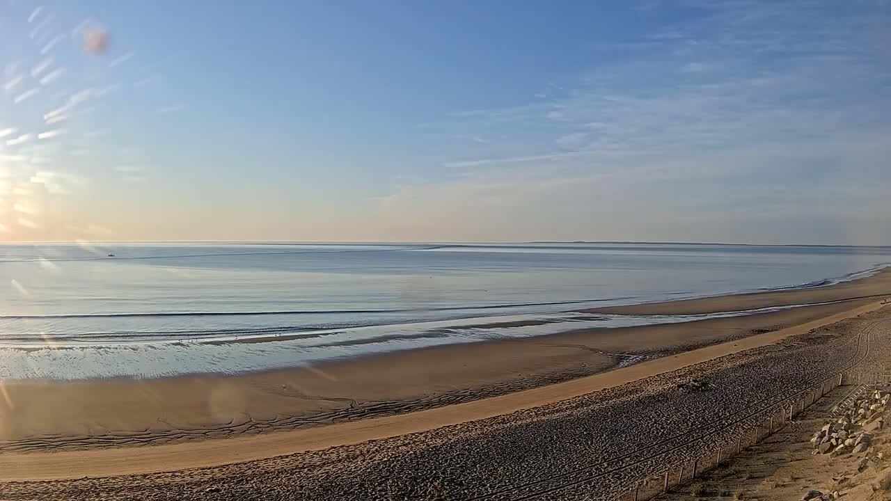 A sandy beach stretches along a calm, sunlit ocean under a mostly clear sky.