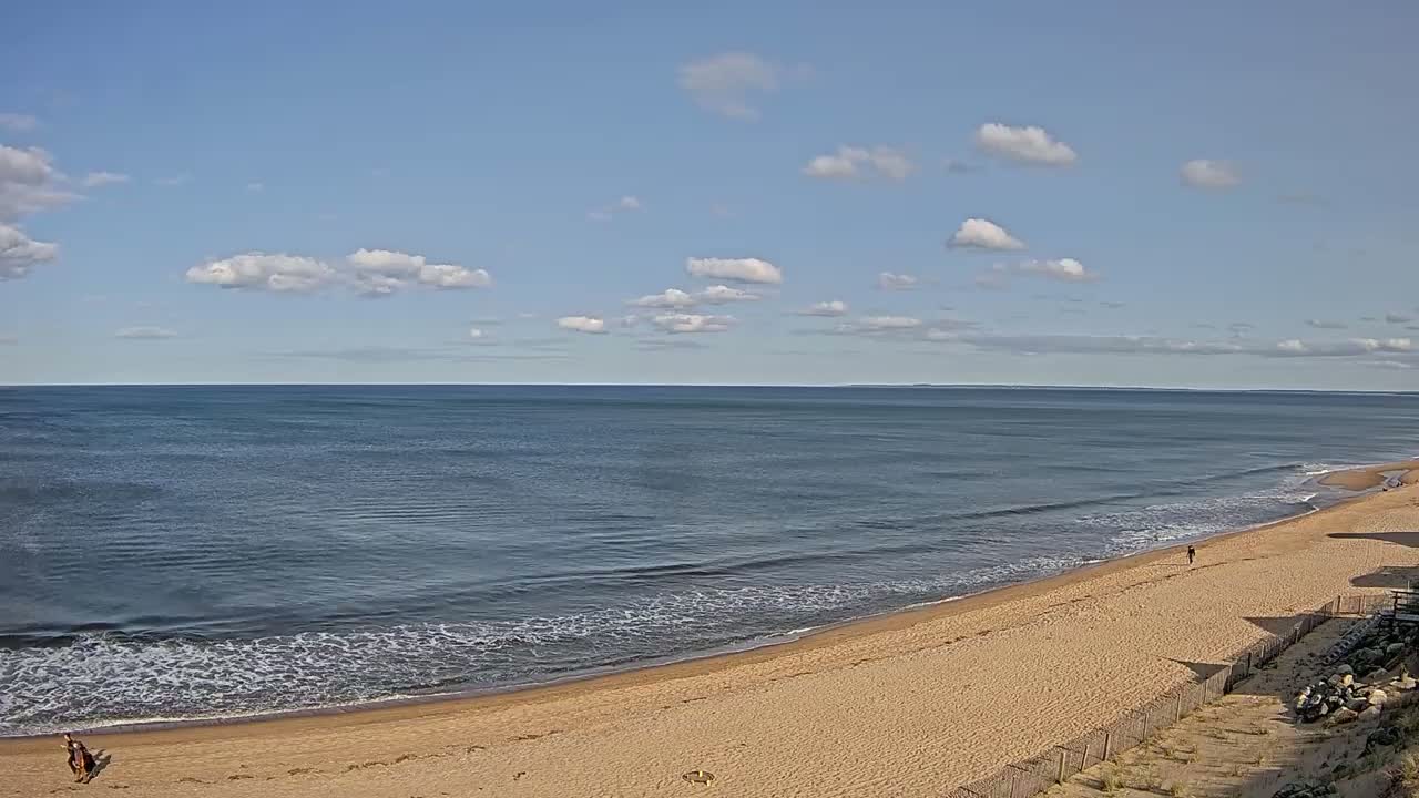 A sandy beach curves gently along a calm ocean under a mostly clear, sunny sky with scattered cumulus clouds.
