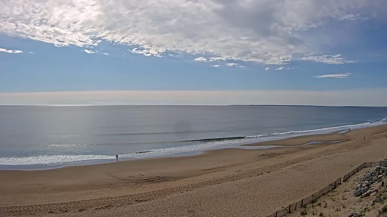 A mostly sunny day shows a long sandy beach with small waves lapping the shore and a few people walking along the waterline.