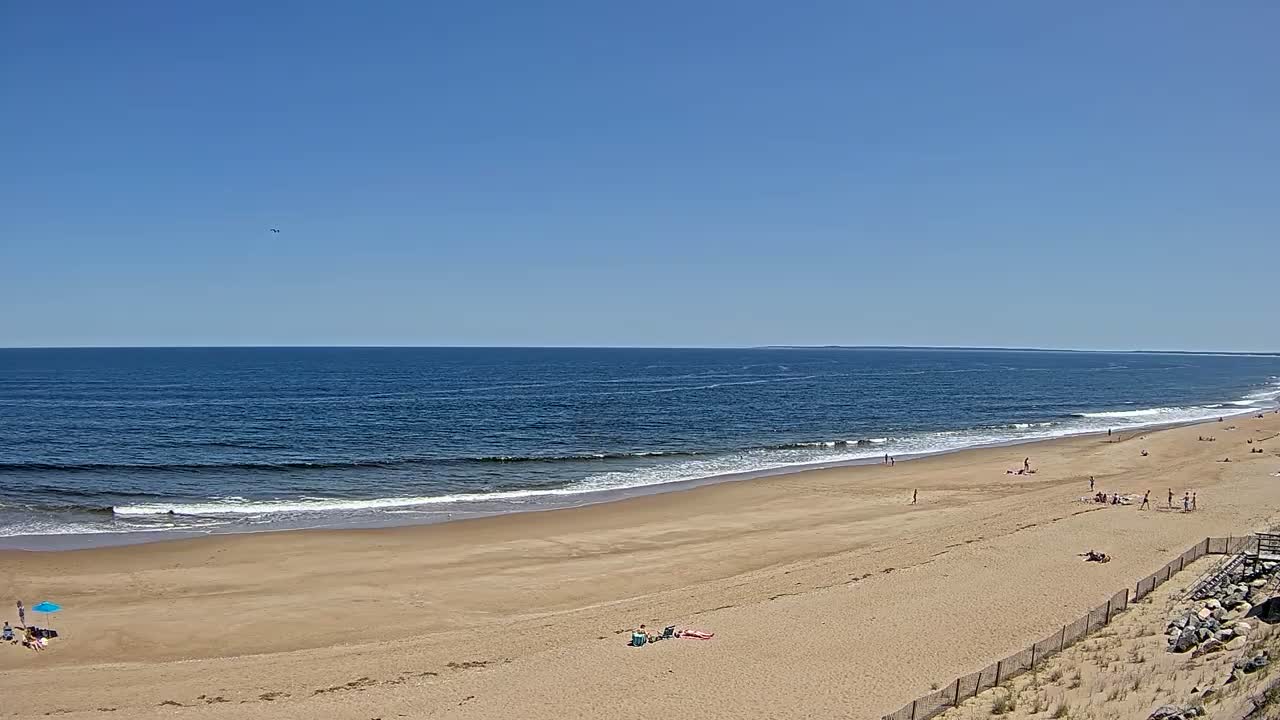 A wide sandy beach stretches along a calm ocean under a clear blue sky, with a few people scattered along the shore.