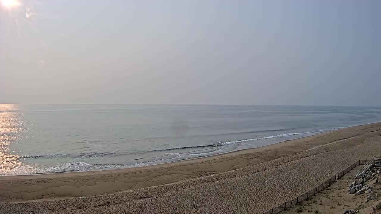 A hazy sunrise illuminates a calm ocean meeting a wide, sandy beach.