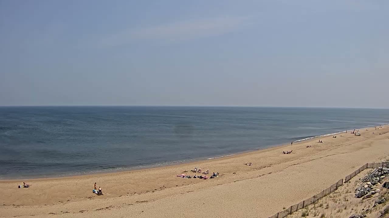 A long sandy beach curves gently along a calm, blue ocean under a clear, bright sky.