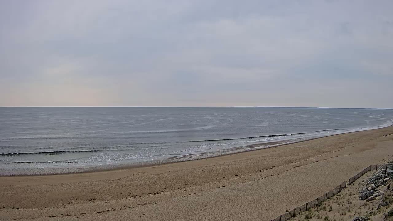 A long, sandy beach slopes gently down to a calm, gray ocean under a cloudy sky.