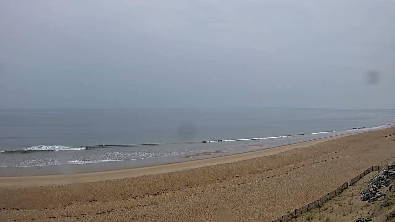 A long, sandy beach slopes gently down to a calm, gray ocean under an overcast sky.