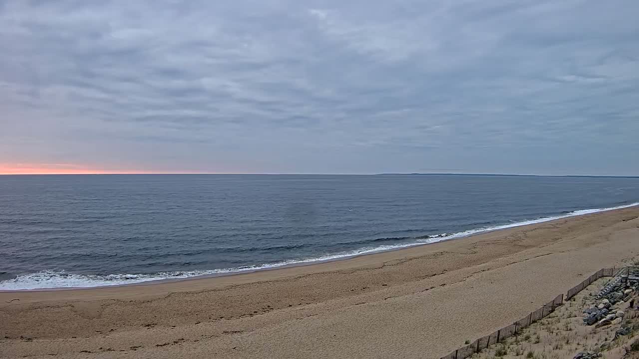 A sandy beach curves gently into a calm ocean under a mostly cloudy sky with a hint of pink on the horizon.