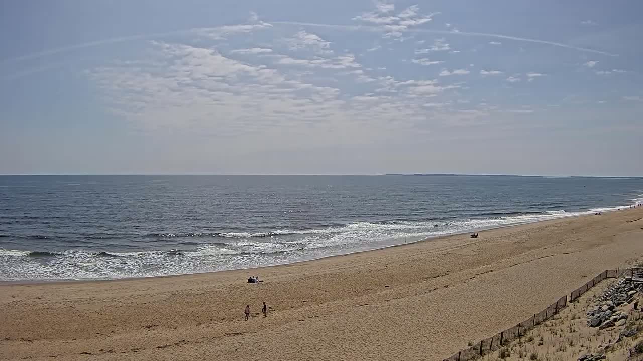 A mostly sunny day shows a wide sandy beach with a few people and gentle waves lapping the shore.