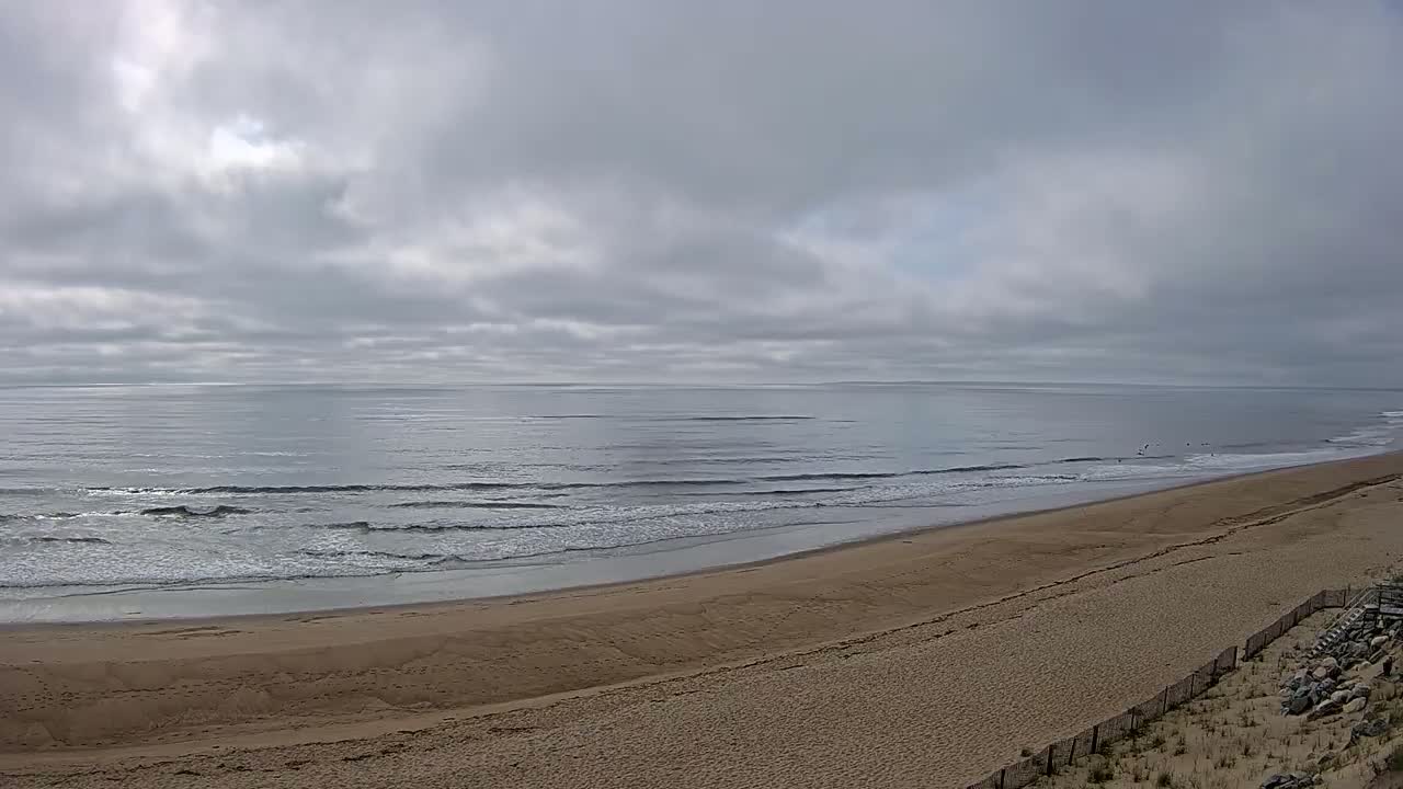 A long, sandy beach slopes down to a calm ocean under a cloudy sky.