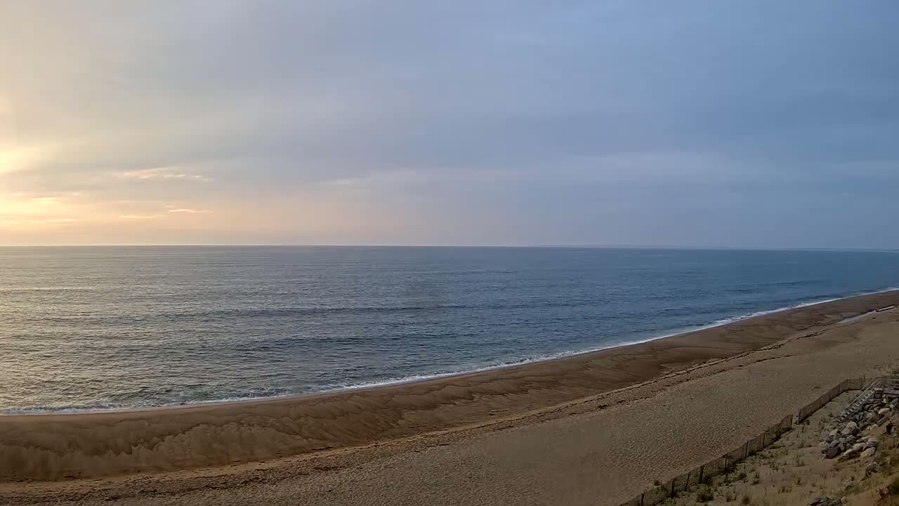 A wide sandy beach curves gently along a calm ocean under a partly cloudy sky at sunrise.