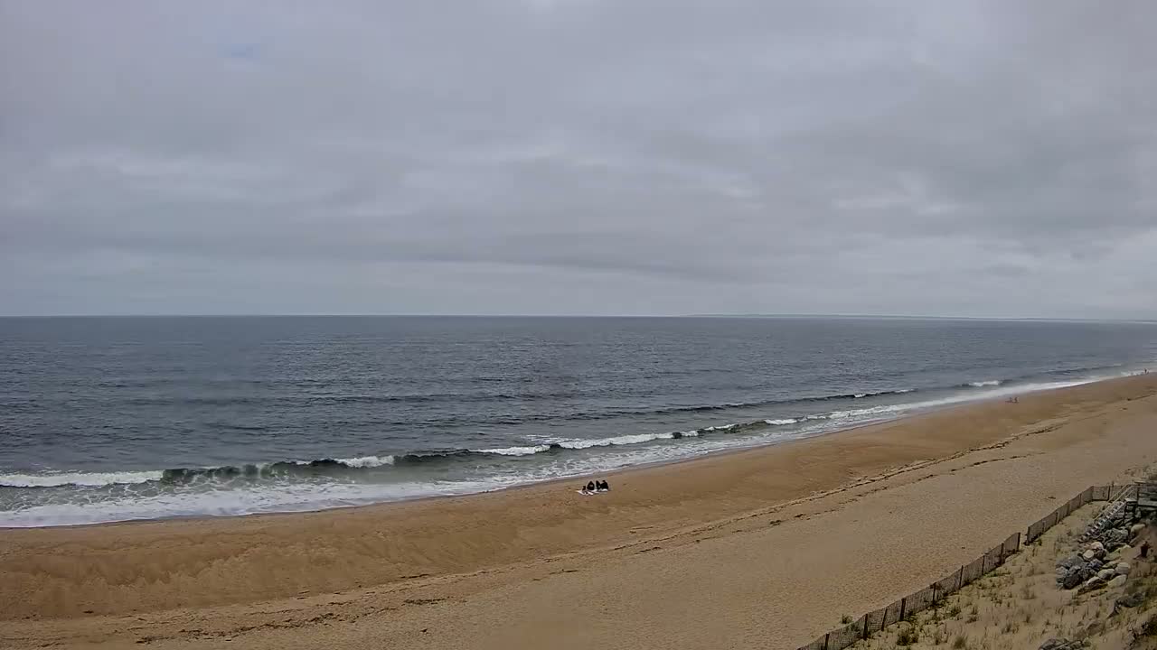A wide sandy beach with small waves under an overcast sky shows a few people sitting on the sand near the water.