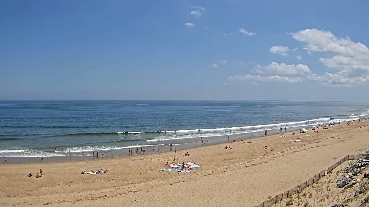 A sandy beach with scattered people is visible under a partly cloudy, sunny sky, with gentle waves rolling onto the shore of a calm ocean.