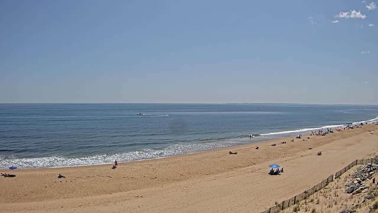 A sandy beach with a few people on it slopes down to a calm ocean under a mostly clear blue sky.