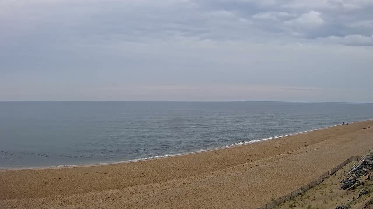 A long sandy beach curves gently towards a calm, grey-blue sea under a mostly cloudy sky.