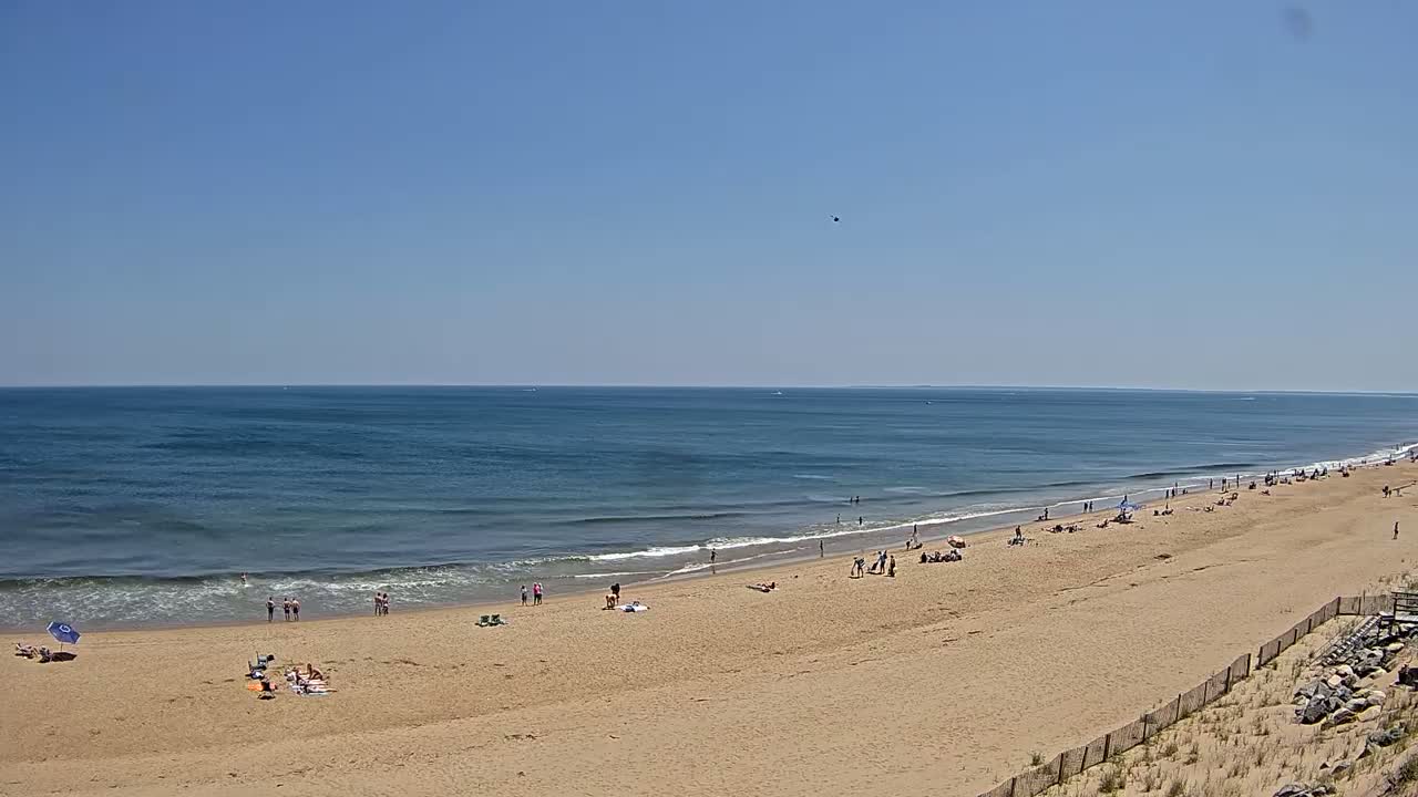 A wide sandy beach with a scattering of people stretches along a calm ocean under a clear blue sky.