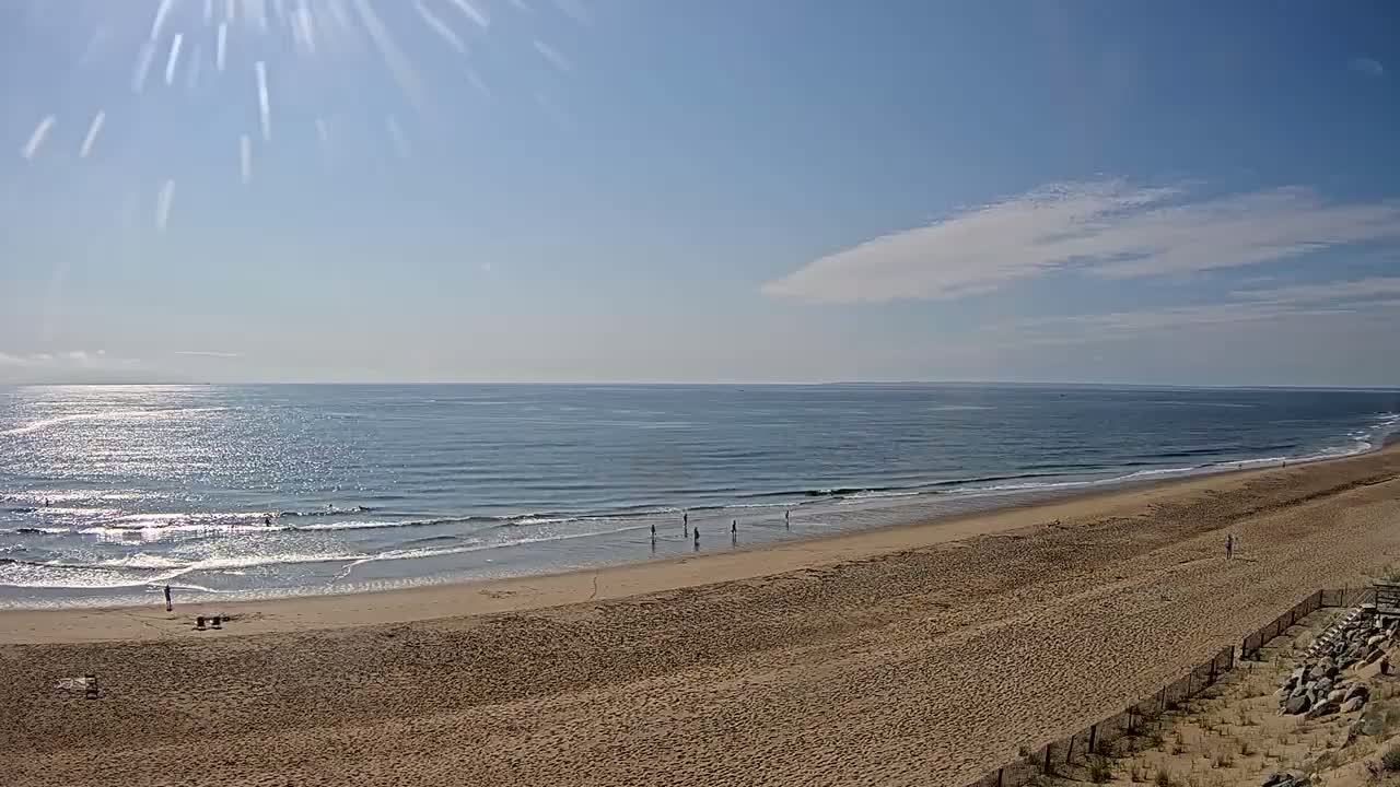 A sandy beach stretches towards a calm ocean under a mostly sunny sky with a few scattered clouds.