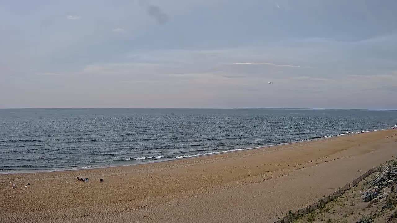 A sandy beach stretches towards a calm ocean under a mostly sunny sky with a few scattered clouds.