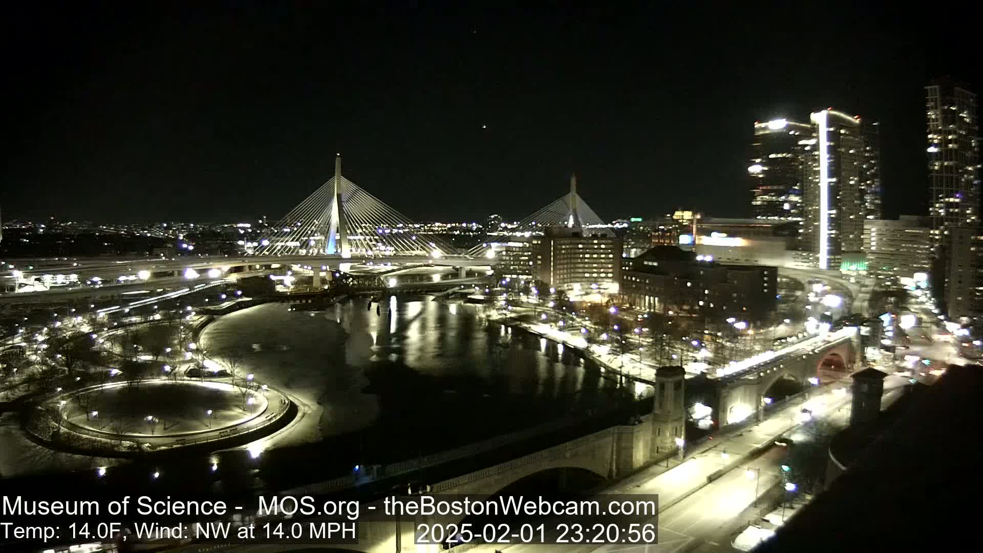 A nighttime view of a city illuminated with lights, showing two cable-stayed bridges spanning a river partially covered in ice, under a clear, cold sky with a northwest wind.