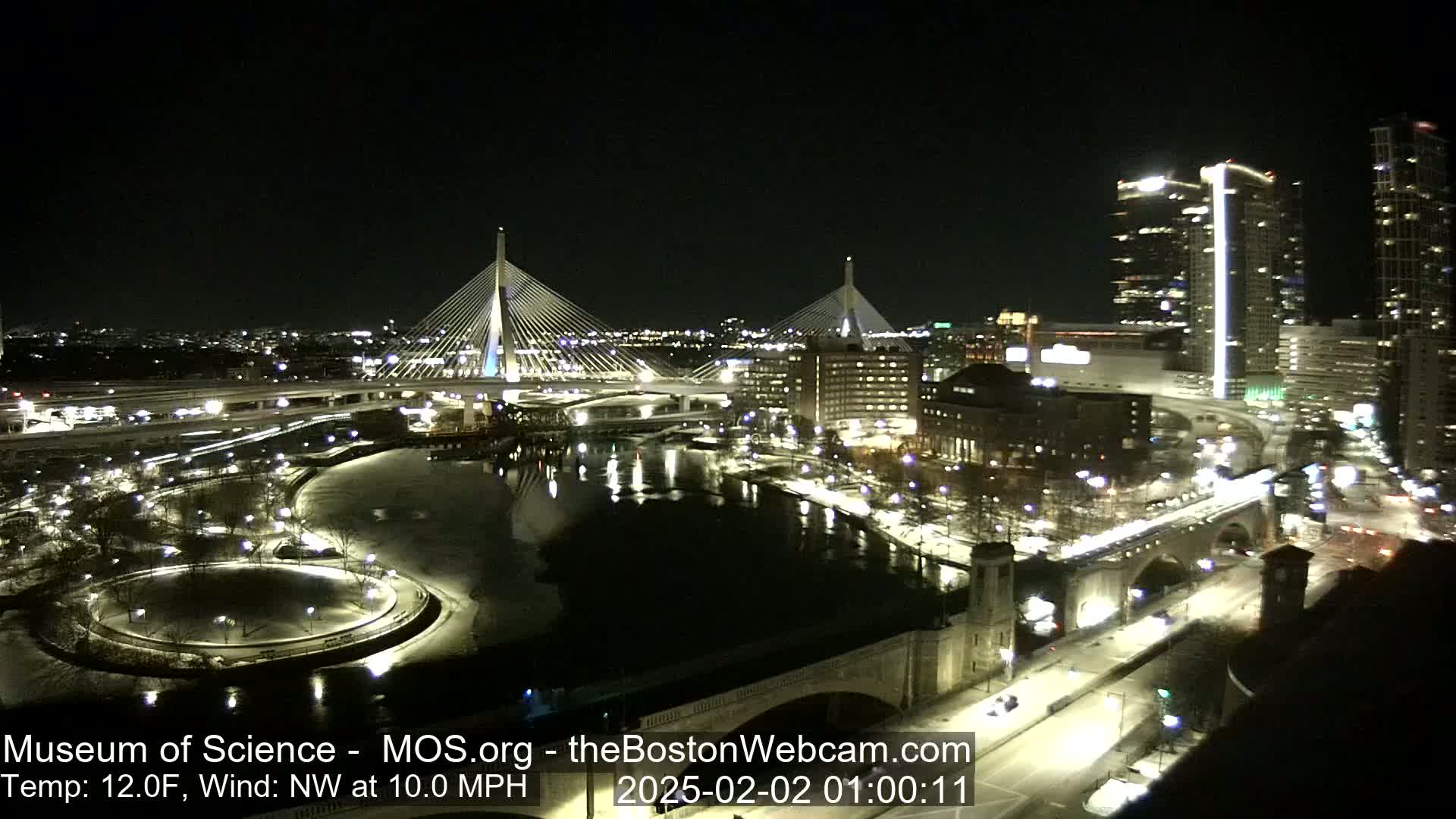 A nighttime, snow-covered cityscape featuring two illuminated cable-stayed bridges spanning a river, with a temperature of 12°F and a northwest wind at 10 mph.