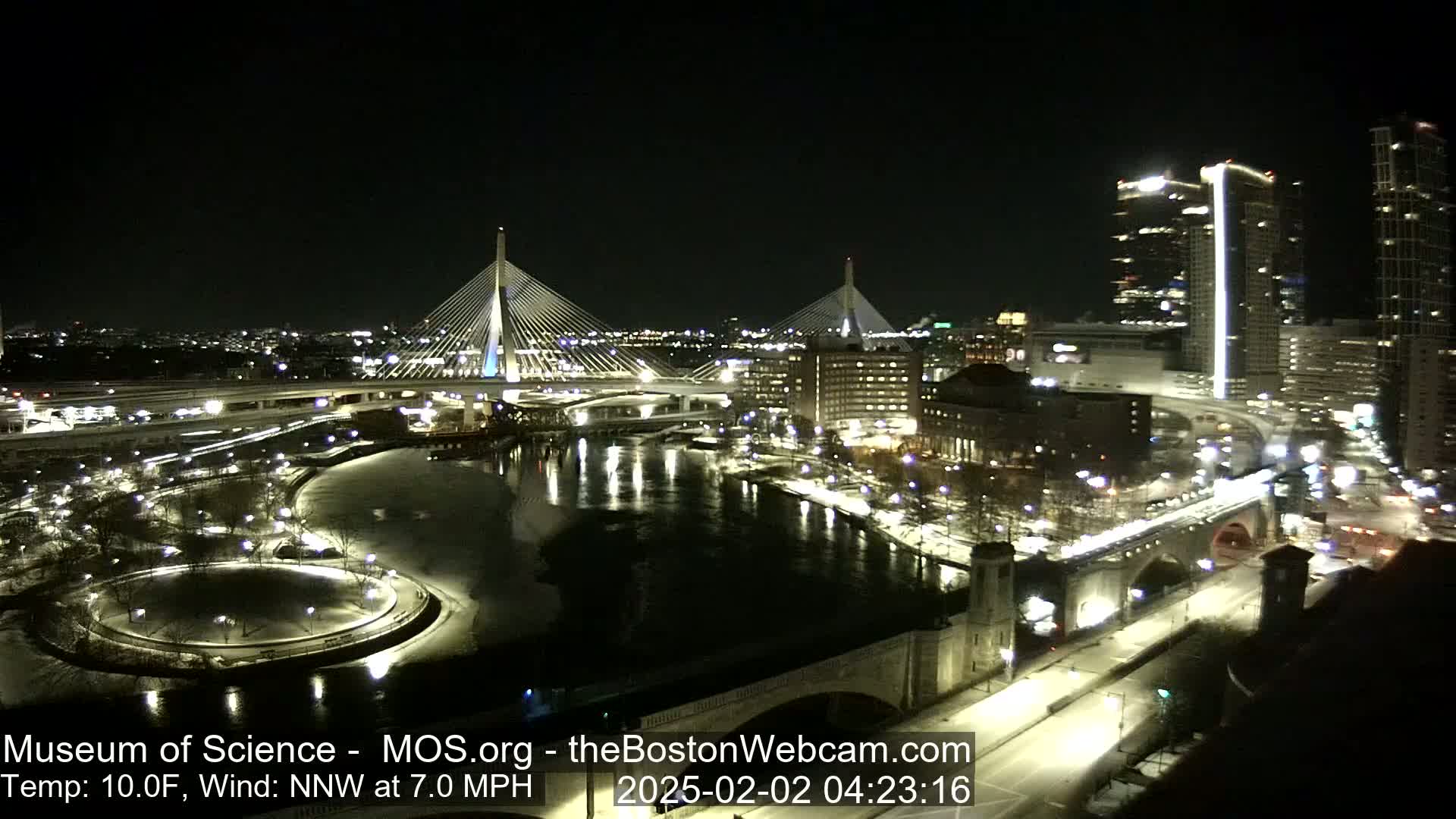 A nighttime view of a city with two cable-stayed bridges spanning a river, under a clear sky with a temperature of 10 degrees Fahrenheit and a 7 mph northwest wind.