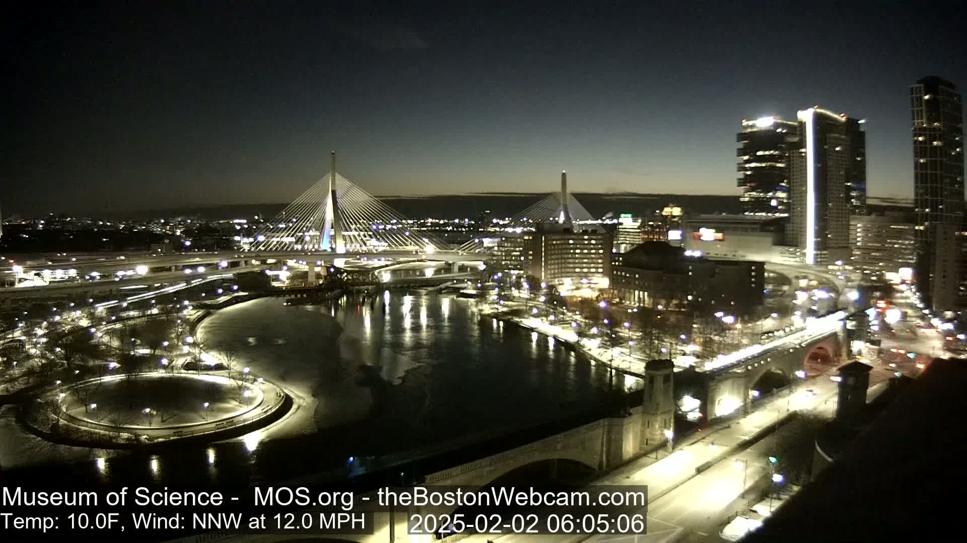 A nighttime cityscape with two cable-stayed bridges over a partially frozen river, under a clear, cold sky.