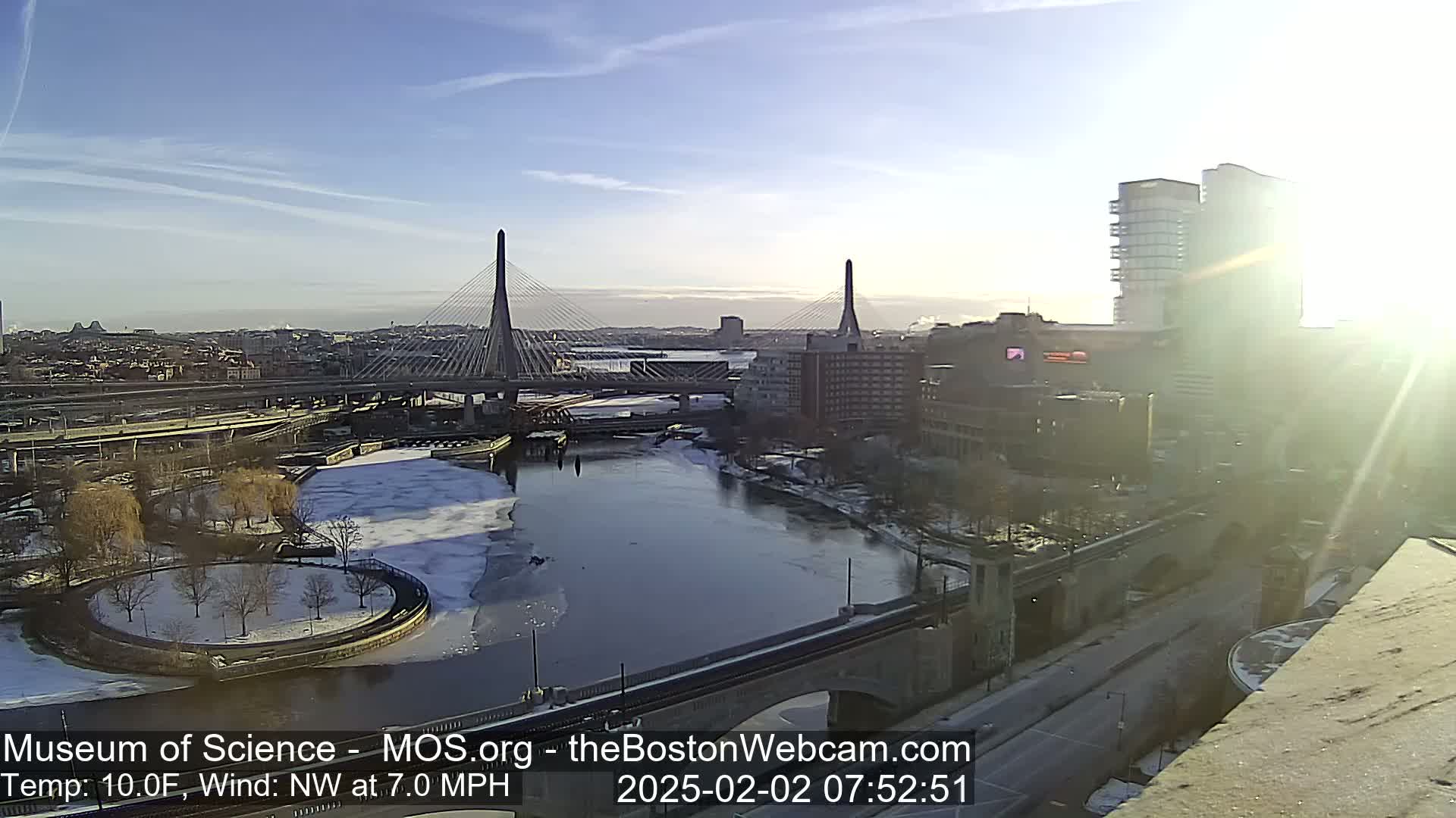 A sunny, cold day shows a cityscape with two cable-stayed bridges spanning a partially frozen river, with some snow on the ground.