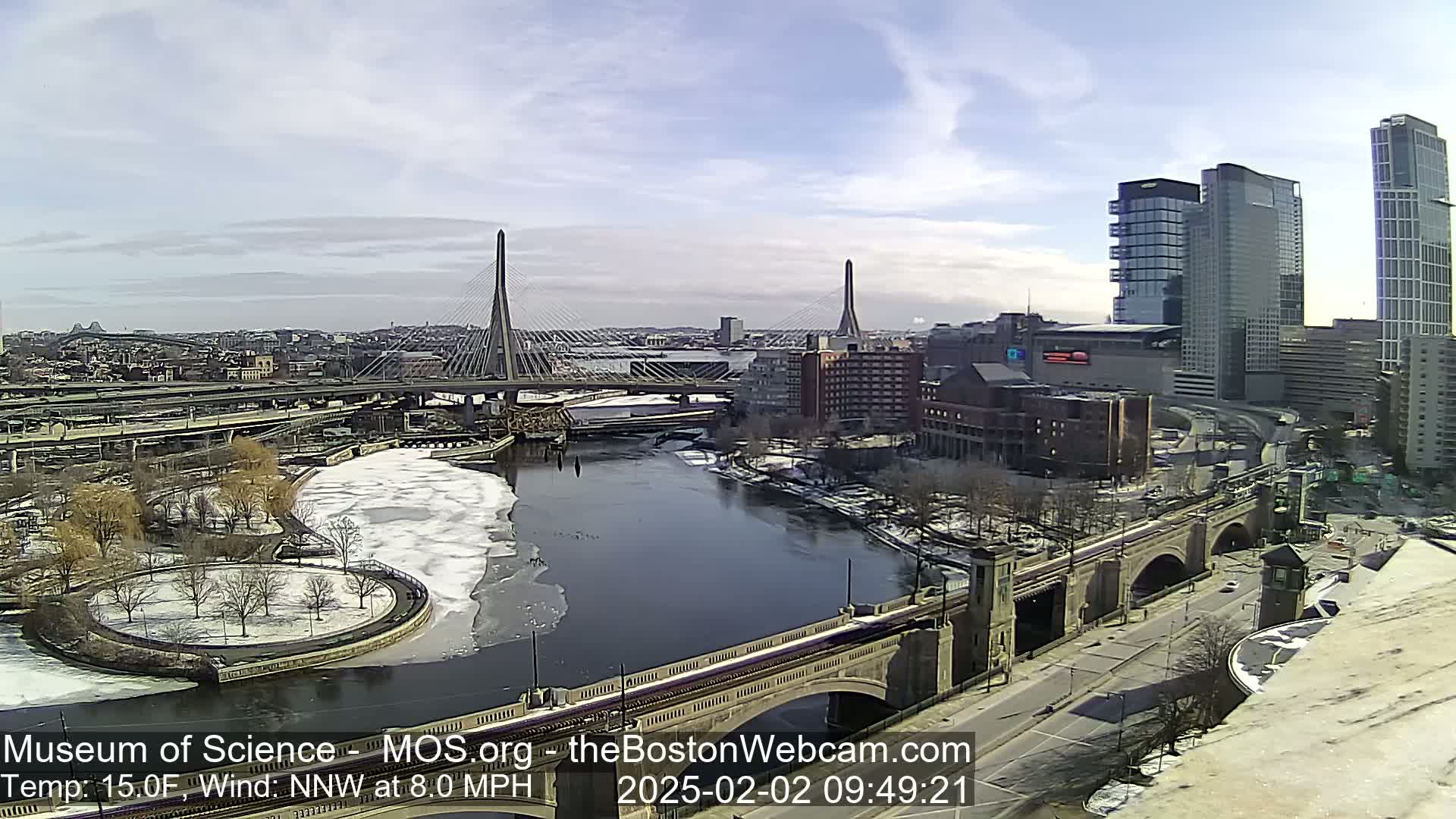 A partly sunny day shows a river partially frozen over, with bridges, city buildings, and roadways visible, under a mostly clear sky.