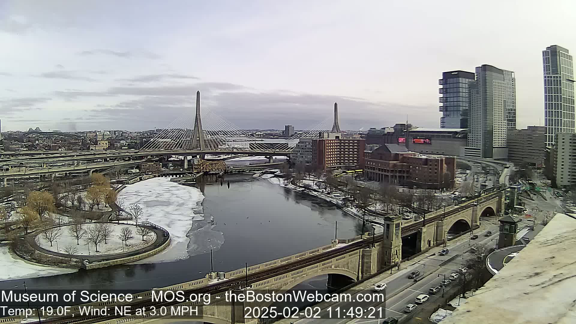A cityscape with two cable-stayed bridges spanning a partially ice-covered river under a cloudy sky.