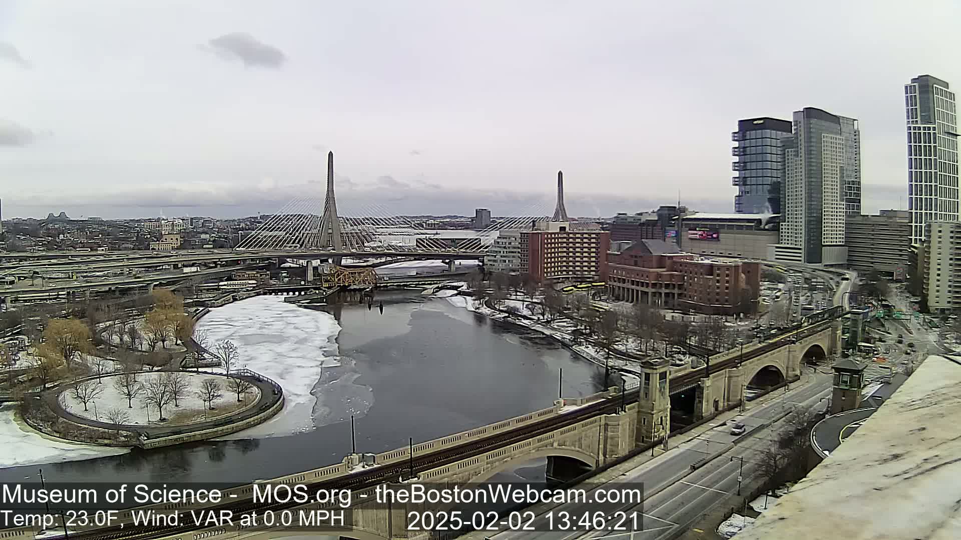 A partly cloudy, cold day shows a city skyline with two cable-stayed bridges spanning a partially ice-covered river.